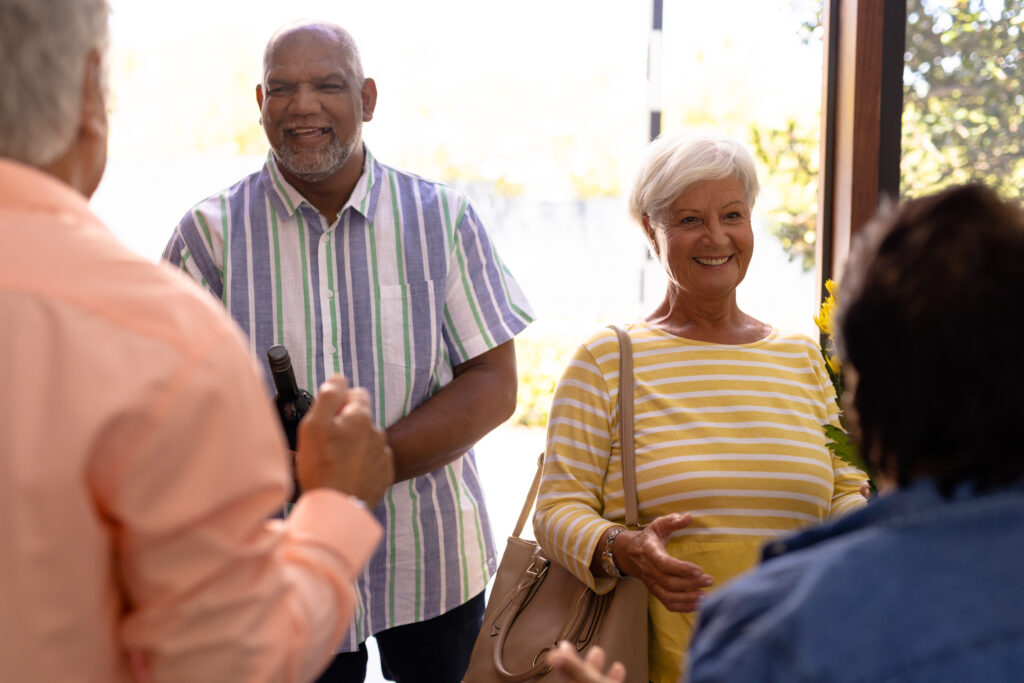 seniors welcoming cheerful friends standing at doorway in nursing home, copy space. Happy, entrance, guest, greeting, unaltered, togetherness, support, assisted living, retirement concept.